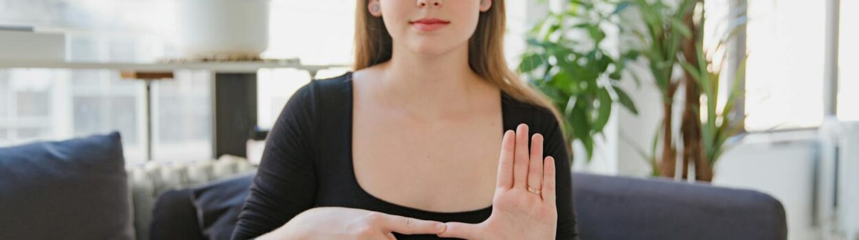 Une femme utilisant la langue des signes est assise sur un canapé dans un salon lumineux entouré de plantes.