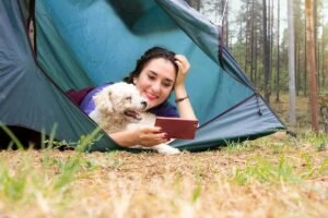 Femme allongée dans une tente bleue avec un petit chien beige, forêt en arrière-plan