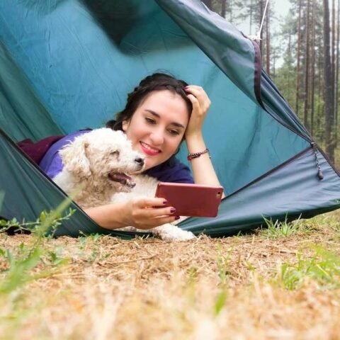 Femme allongée dans une tente bleue avec un petit chien beige, forêt en arrière-plan