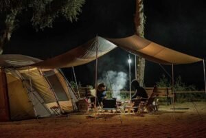 Campement de nuit avec tente, auvent tendu et deux personnes près d'un foyer fumant sous les arbres