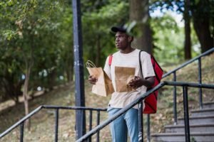 Homme noir portant un sac à dos rouge, descendant des marches en métal, tenant deux sacs en papier