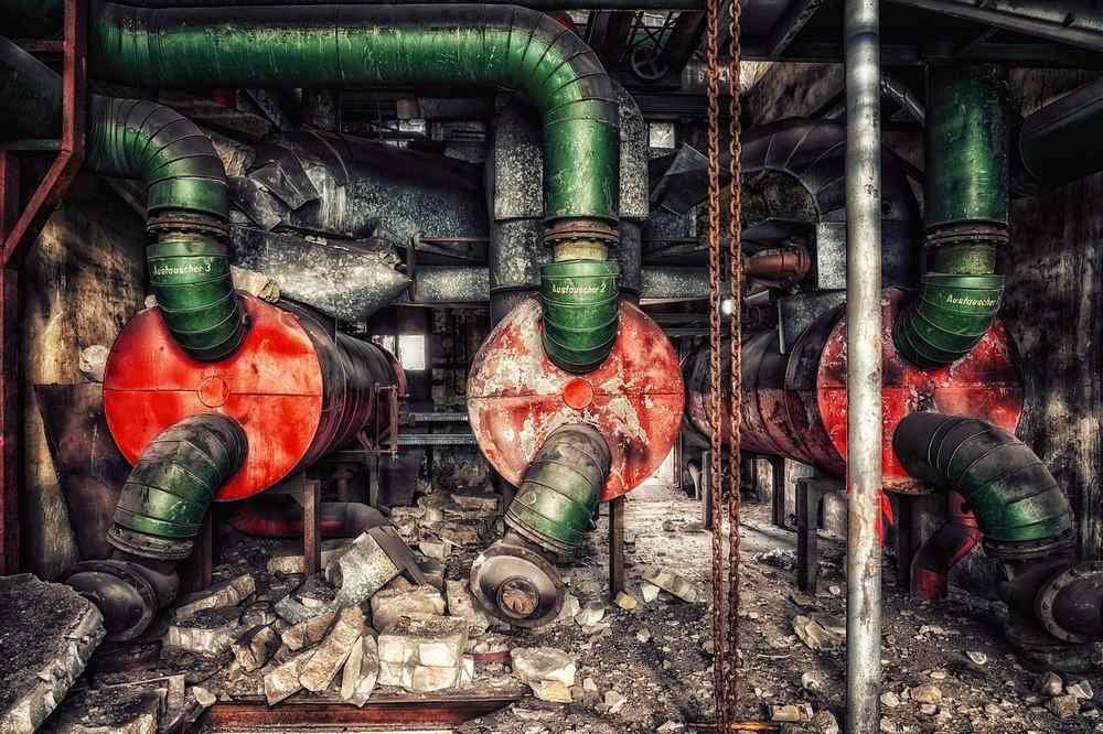 Tuyaux verts, cuves rouges rouillées, débris au sol dans une usine abandonnée