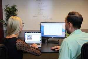 Deux collaborateurs assis à un bureau regardent ensemble un ordinateur portable et un écran de bureau