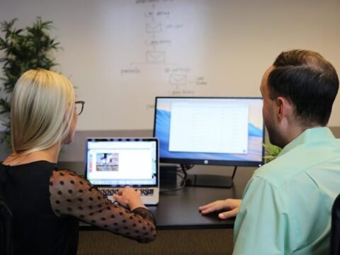 Deux collaborateurs assis à un bureau regardent ensemble un ordinateur portable et un écran de bureau