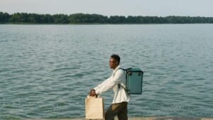 Homme noir marchant avec deux sacs près d'un lac, arbres en arrière-plan