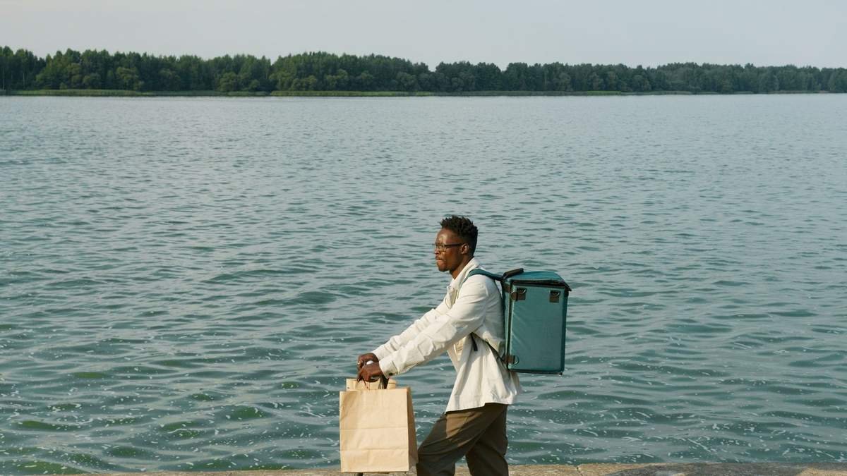 Homme noir marchant avec deux sacs près d'un lac, arbres en arrière-plan