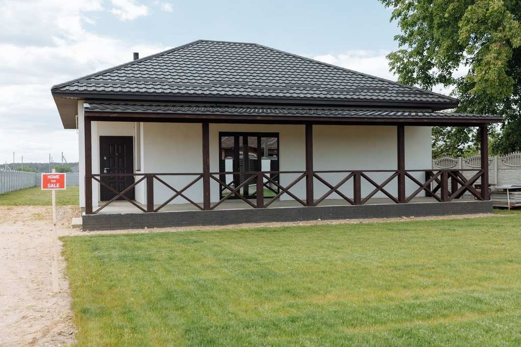 Maison blanche avec balcon couvert et porte d'entrée, panneau signalétique rouge, pelouse verte, ciel partiellement nuageux Maison blanche avec balcon couvert et porte d'entrée, panneau signalétique rouge, pelouse verte, ciel partiellement nuageux